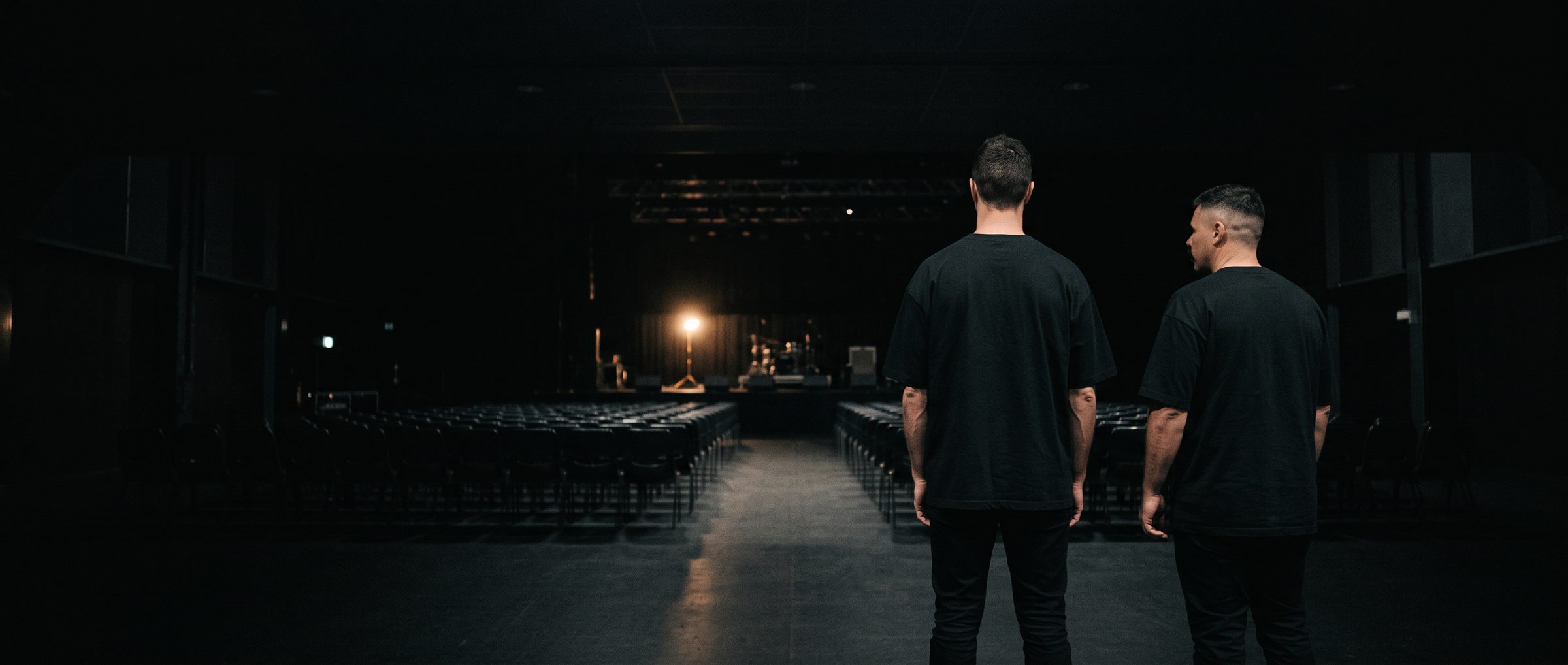 Two men in black T-shirts seen from behind, standing in the aisle of a dark, empty auditorium facing a lit stage and rows of chairs